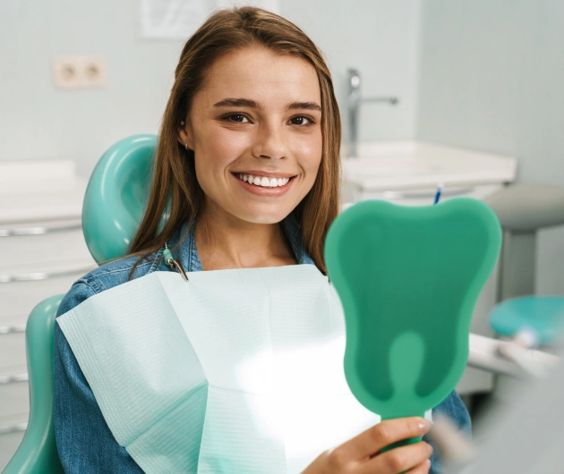 Smiling female dental patient in treatment chair holding model teeth at dental office.