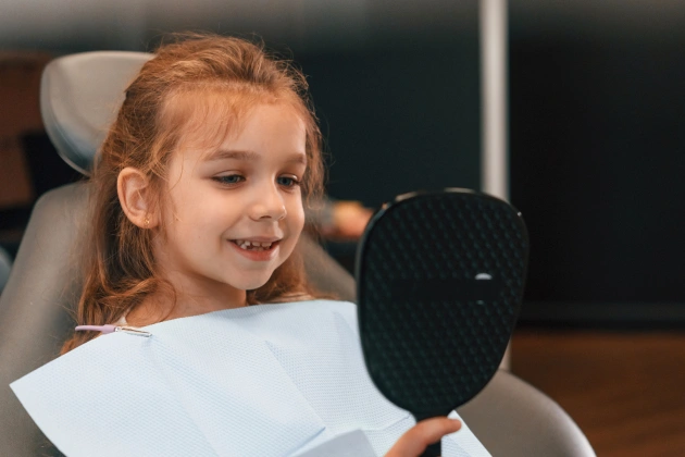 Young girl smiling at dental office holding mirror during examination.