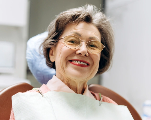 Smiling woman wearing glasses in dental office setting, appearing happy and relaxed in chair.