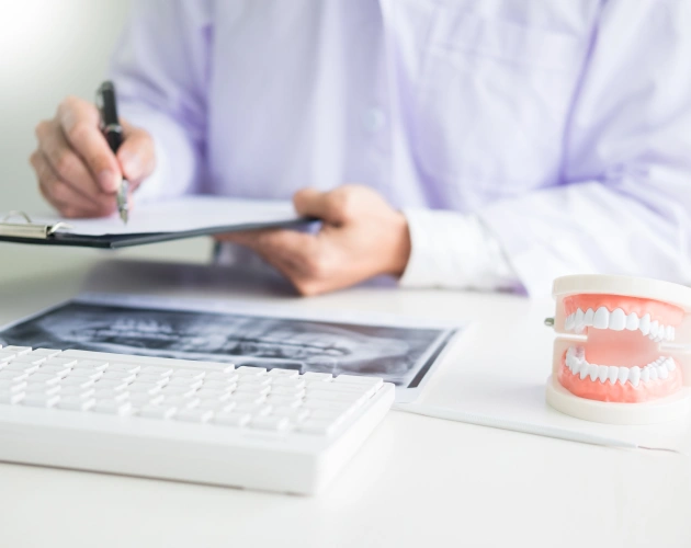 Dentist writing notes at desk with dental model and laptop in clinical office setting.