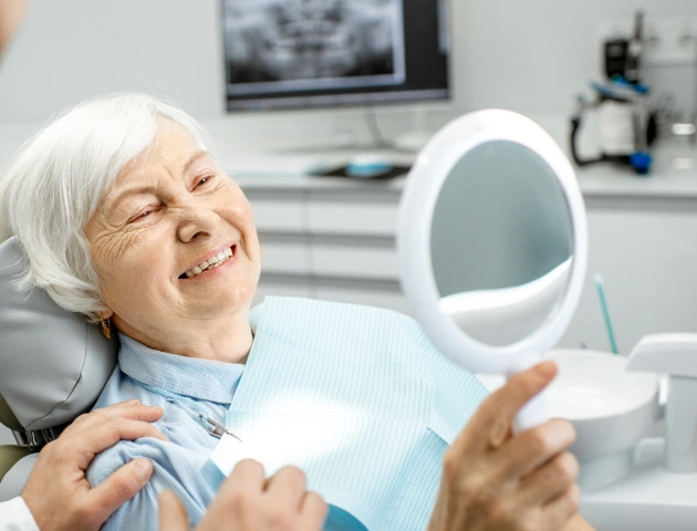 Senior woman examining her smile in mirror at dental office during checkup appointment