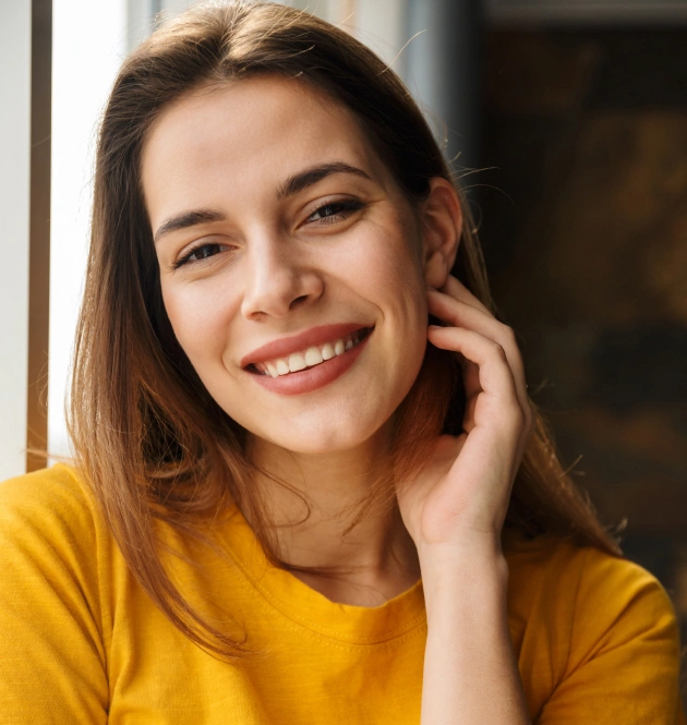 Smiling woman in yellow shirt touches face, warm indoor setting, dental care portrait.
