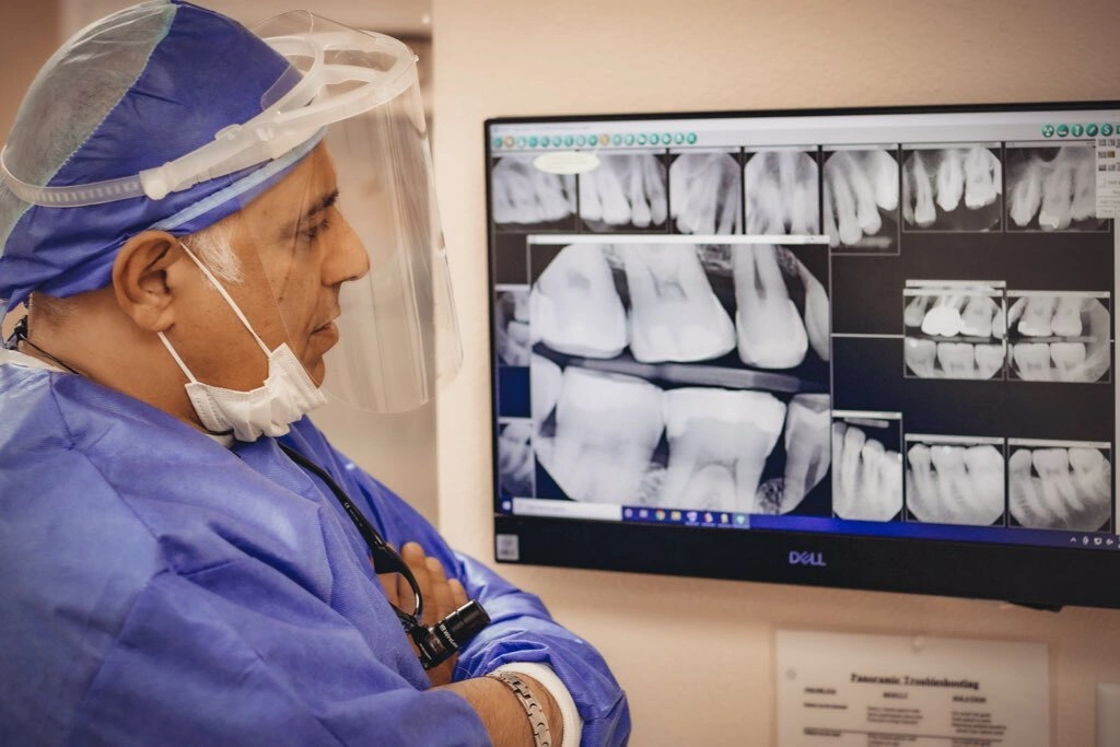 Dentist in surgical attire examining dental X-rays on monitor in clinical setting.