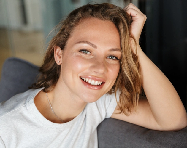 Smiling woman in white shirt relaxing on gray couch in casual indoor setting.