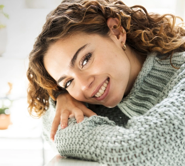 Woman smiling warmly while hugging herself in cozy sweater, dental care setting.