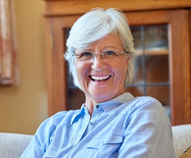 Smiling older woman with white hair wearing glasses and blue shirt in dental office interior.