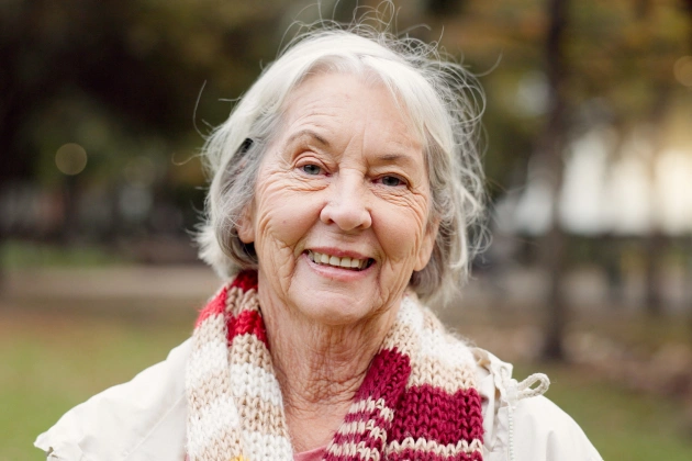 Smiling elderly woman wearing white sweater and red scarf outdoors in park setting.