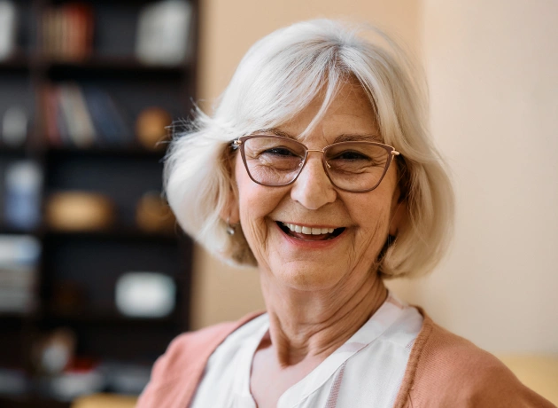 Smiling older woman with white hair and glasses in dental office setting with bookshelves behind her.
