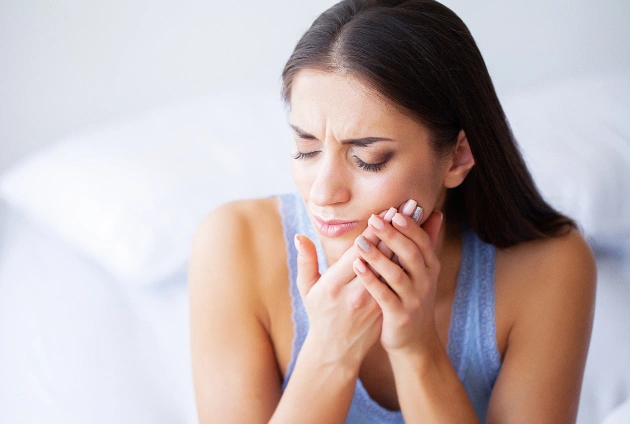 Woman holding face in pain while sitting on bed, expressing dental discomfort or toothache.