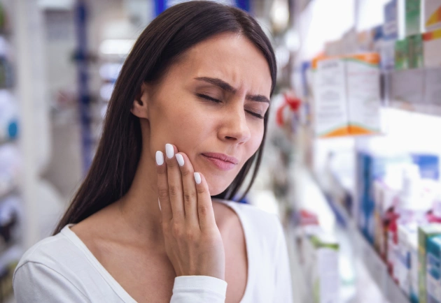 Woman experiencing tooth pain in pharmacy, holding cheek with concerned expression near medication shelves.