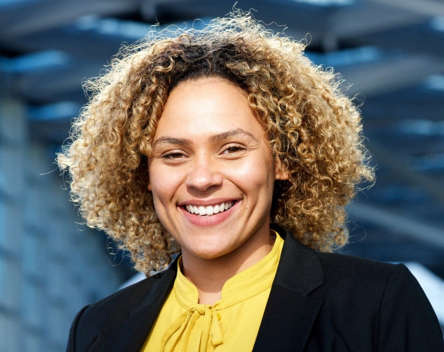 Woman with curly blonde hair smiling at camera wearing yellow top and black blazer.