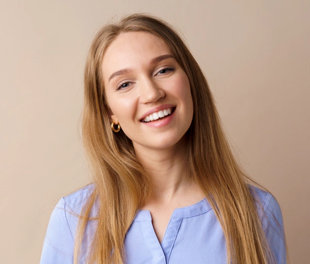 Young woman smiling at camera wearing light blue top against neutral beige background