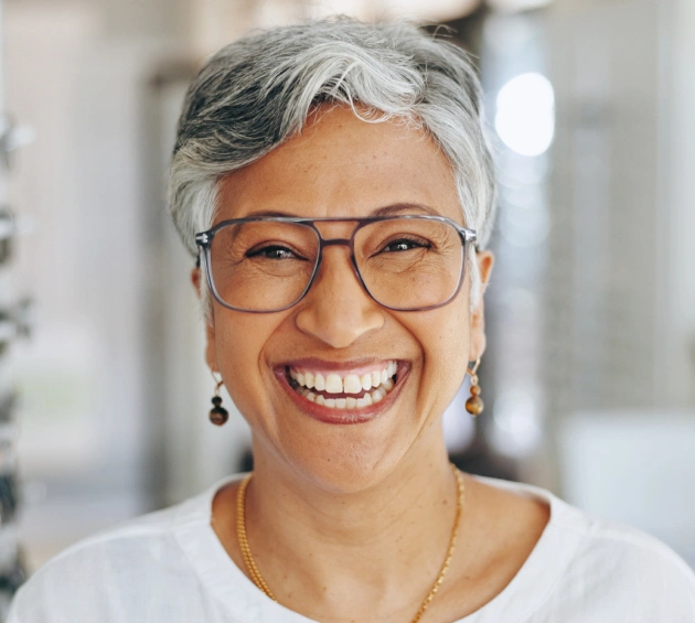 Smiling woman with gray hair and glasses in dental office setting wearing white shirt.