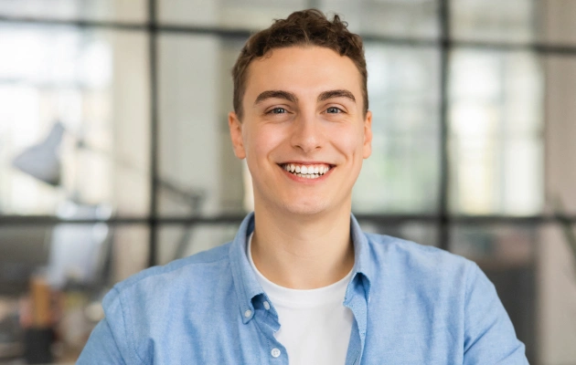 Smiling male dental professional in blue shirt, modern office setting with windows behind him.