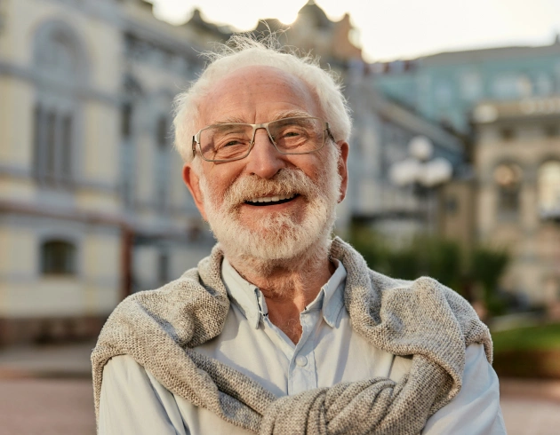 Smiling older man with glasses wearing vest in outdoor urban setting near buildings.