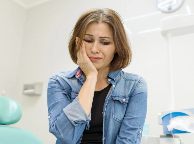 Woman in denim shirt with hand to face, contemplative expression, dental office setting with equipment visible.