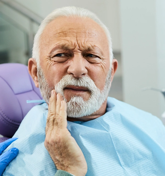 Elderly man in dental bib touching cheek, appearing uncomfortable in dental office chair.