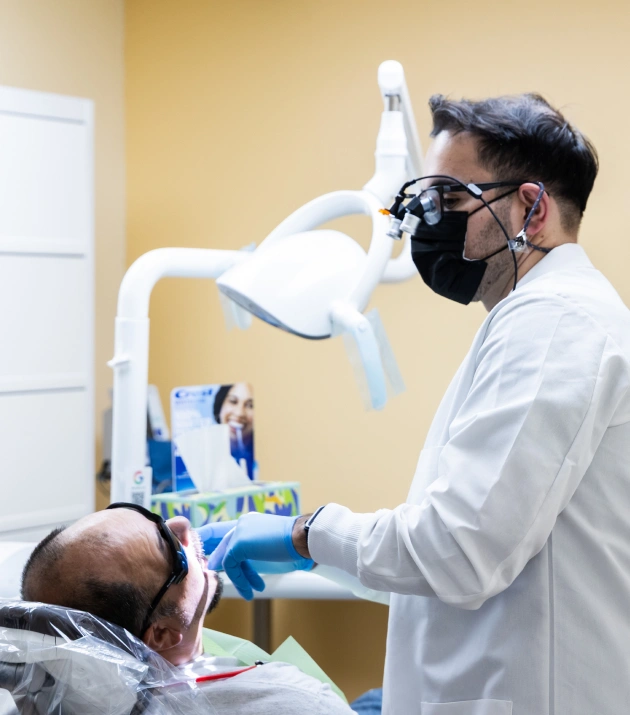 Dentist in mask and loupes examining patient in dental chair under overhead light.