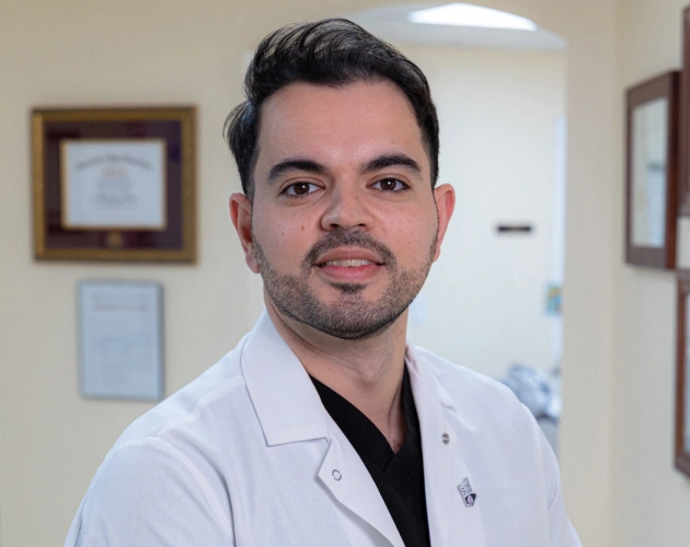 Male dentist in white coat and black shirt poses in dental office setting.