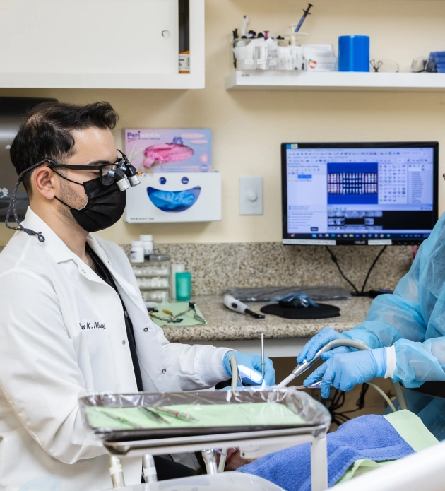 Dental professional in mask and gloves examines patient in modern dental office with equipment and monitors.