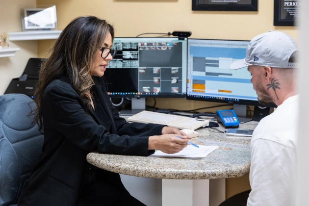 Dental receptionist handing paperwork to male patient at front desk with computers visible.