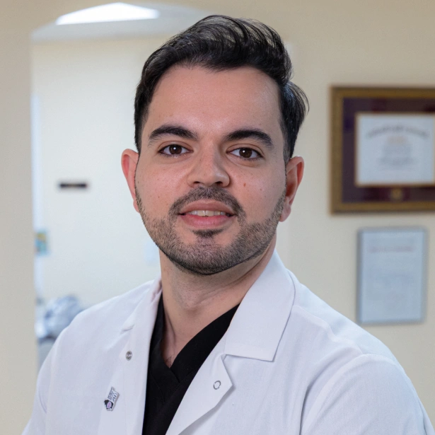 Male dentist in white coat and black shirt smiling at camera in dental office setting.