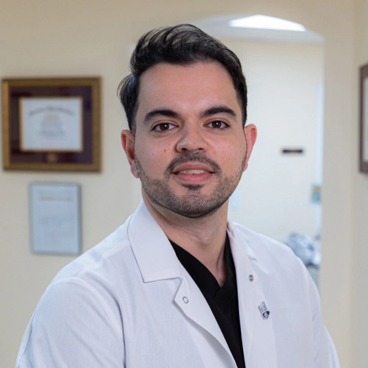 Male dentist in white coat and black shirt standing in modern dental office.